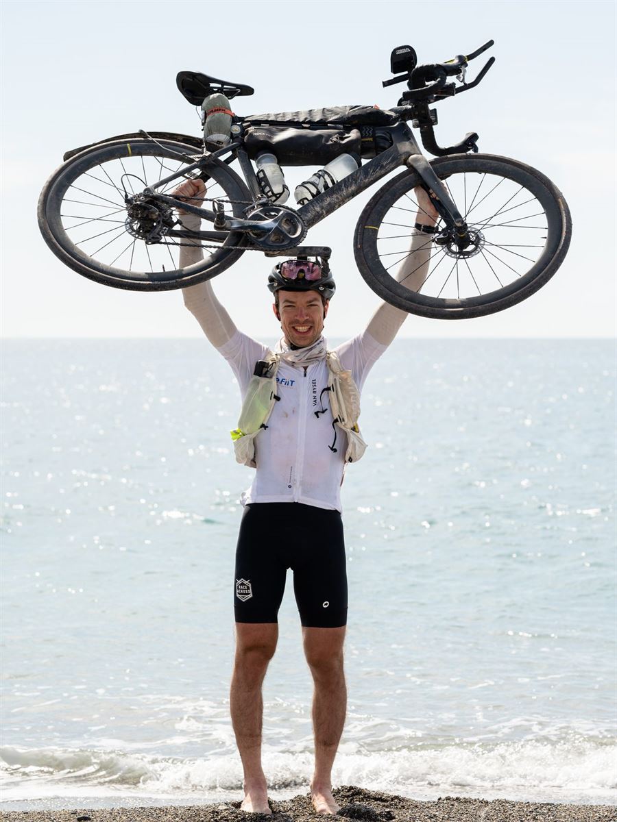 Bastien, bras levés, brandissant son vélo de bikepacking au bord de la mer.